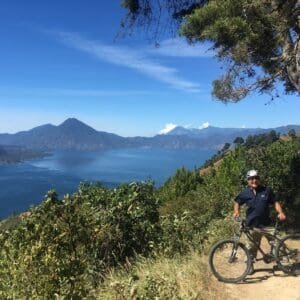 Man with bike stands behind a postcard view of Lake Atitlan from anove.