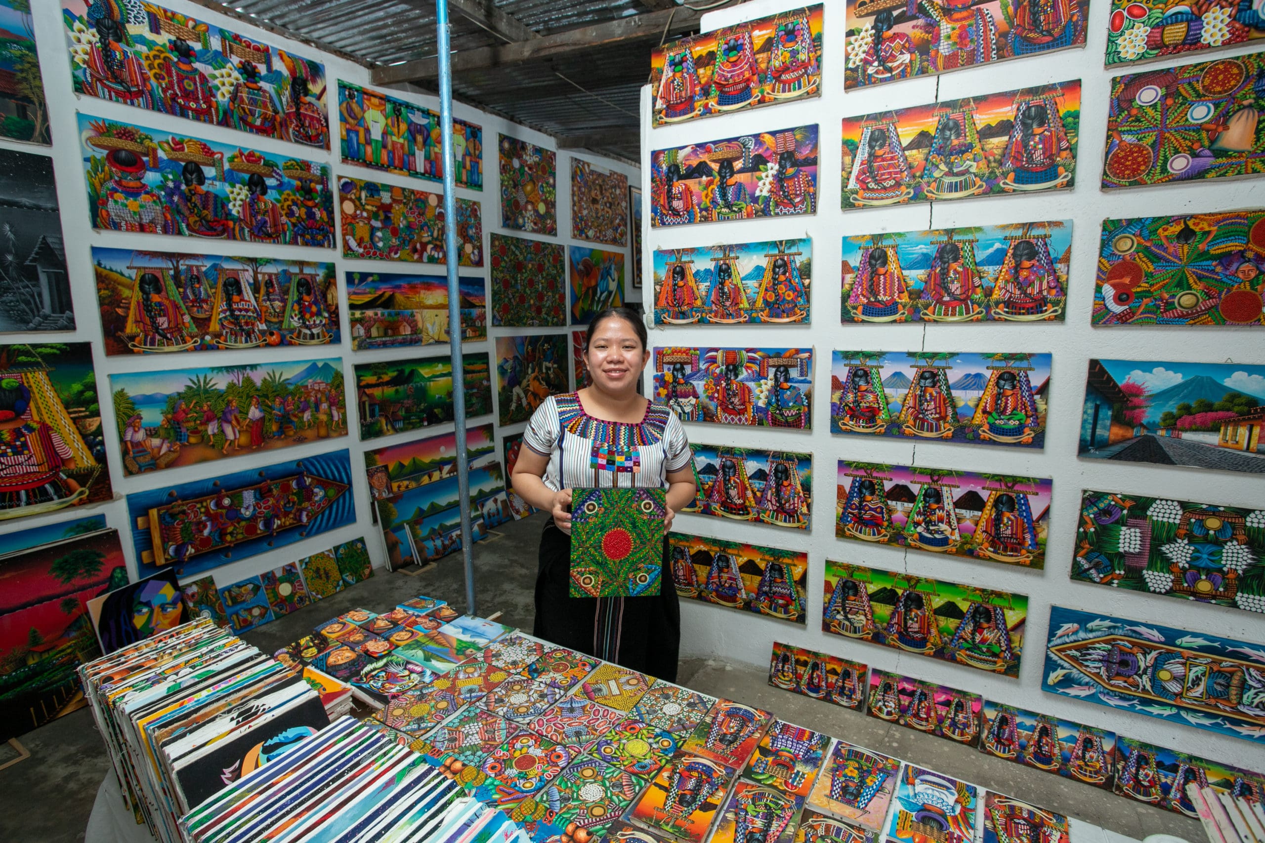 Woman holds artwork in front of wall filled with paintings.
